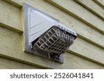 The exterior of a bright yellow house wall with a white dryer vent exposes the air vent which is clogged with hair, wood, and dirt in the mesh causing a fire hazard. The plastic unit guard is dirty.