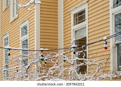 The Exterior Of A Building With Colorful Christmas Lights Hangs In The Garden Of A Yellow Wooden Clapboard House With Cream Color Trim. The Tree In The Yard Is Covered In White Wintery Snow. 