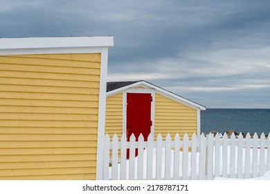 The exterior of colorful wooden sheds with a red door overlooking the blue ocean and cloudy sky. A white picket fence in the yard with white snow piled up next to the beach hut style building. - Powered by Shutterstock