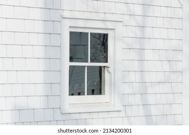 The exterior corner of a vintage wooden building with a closed glass single hung window. The wall is covered in white shingles. There's a yellow wooden building in the background with white trim. 