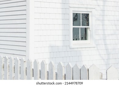 The exterior corner of a vintage wooden building with a closed glass single hung window. The wall is covered in white shingles and a shadow. There's a white picket fence attached to the building. 