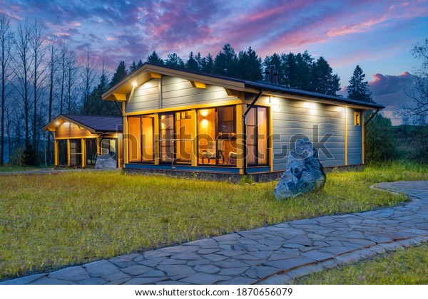 Exterior of a country cottage. Small houses on\
background of forest. Scandinavian style wooden houses. Lawn and\
stone path next to cottage. Architectural design of a Scandinavian\
house. Evening time