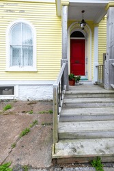 The Exterior Entrance To A Bright Yellow House With Horizontal Wooden Clapboard Siding, Arched Glass Window, Red Metal Door, Flower Pot, Wooden Front Steps, And White Trim Around The Door And Window.