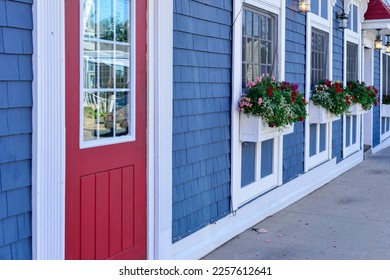 The exterior entrance to a shop with a bright red door and glass window. There are three windows in a row on the colorful blue cedar shake wall with white trim. The flower boxes have summer flowers. - Powered by Shutterstock