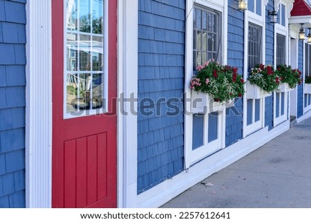 The exterior entrance to a shop with a bright red door and glass window. There are three windows in a row on the colorful blue cedar shake wall with white trim. The flower boxes have summer flowers.