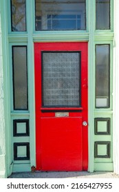 The exterior facade of a vintage shop with an orange wooden door with a glass window, mailslot, and door handle.The door is on a lime green building with black trim. There's a transom window over door