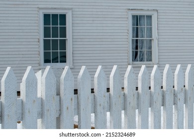 The exterior front yard of a vintage wooden building with closed glass single hung windows. The wall is covered in white wood shingles. There's a white picket fence in front of the building. 