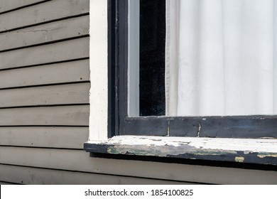 An Exterior Grey Wood Wall Of A House With A Wooden Window Frame, Solid Glass And White Curtain. The Clapboard Board Is Narrow And The Trim Around The Black Window Is Worn With The Paint Peeling.