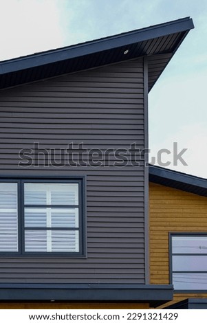 The exterior of a grey and yellow colored modern pvc or vinyl siding corner walls of a house. The flat roof is sloped with a blue colored painted eave. The windows have multiple glass panes with blue.