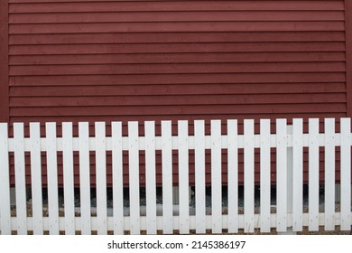 Exterior Horizontal Clapboard Wooden Bright Red Wall With The Sun Shining On It. There's A White Wooden Picket Fence In The Foreground. The Narrow Boards Are Textured Wood In A Linear Pattern.