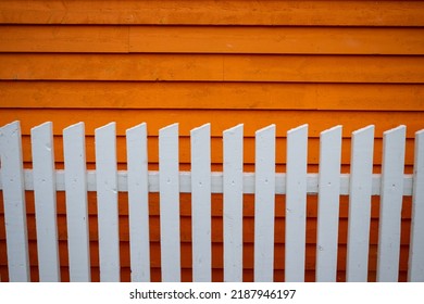 Exterior Horizontal Clapboard Wooden Bright Orange Wall With The Sun Shining On It. There's A White Wooden Picket Fence In The Foreground. The Narrow Boards Are Textured Wood In A Linear Pattern.