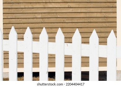 Exterior Horizontal Clapboard Wooden Tan Colored Wall With The Sun Shining On It. There's A White Wooden Picket Fence In The Foreground. The Narrow Boards Are Textured Wood In A Linear Pattern.