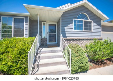 Exterior Of A House With Bricks And Vinyl Sidings And Glass Windows. There Are Plants On Either Side Of The Porch Stairs And Storm Door With Window Panel On The Side.