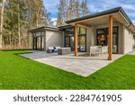 Exterior image of a contemporary home with flat roof and brown trim and lush green grass blue sky forest in the background furnished patio for outdoor dining and lounging