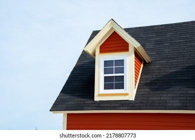 The exterior of a red wooden clapboard building with a steep black shingled roof. In the attic of the house is a closed glass window and dormer with white trim. The background is blue cloudy sky.