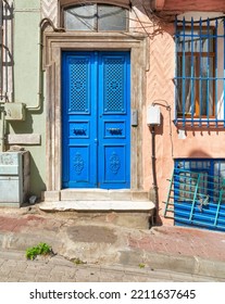 Exterior Shot Of Blue Painted Decorated Metal Door Beside Wrought Iron Window, In A Colorful Stone Wall Painted In Orange And Green, On City Street