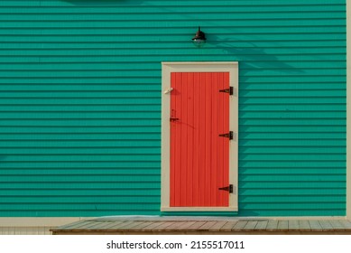 An exterior single traditional wooden shutter door with cream color trim, black hinges, and vintage latch on a vibrant teal green color exterior wall of a house with a light fixture over the door. 
