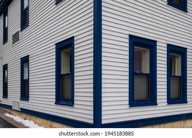 The exterior of a stark white cape cod clapboard horizontal wooden board siding wall with multiple blue trim double hung windows. The corner of the residential building sits on a concrete footing.
