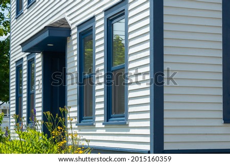 The exterior of a stark white cape cod clapboard horizontal wooden board siding wall with multiple blue trim double hung windows. There are vibrant green flowers in front of the vintage windows. 