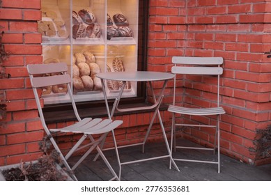 Exterior view of an empty small cafe patio setup. Features a round metal table and two folding chairs positioned against a red brick wall. Adjacent window displays freshly baked bread and