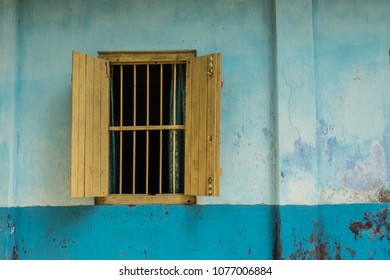 Exterior view of a yellow window with bars and no glass, blue curtains and open yellow shutters in a two-tone blue wall. - Powered by Shutterstock