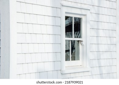 The exterior of a vintage building is covered in white cedar wood shingles. There's a small single hung glass window in the center with the reflection of a white picket fence and tree in the window. 