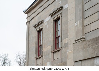 The Exterior Of A Vintage Limestone Block Wall With Multiple Windows. The Tall Government Building Has A Blue Sky With Clouds In The Background. The Trim Around The Double Hung Windows Is Red Wood