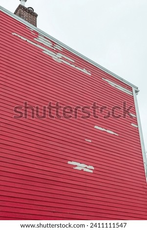 The exterior wall of a bright red wooden house. The multi story building's paint surface is peeling from water and wind damage. The horizontal wood boards are textured timber.There's a brick chimney.