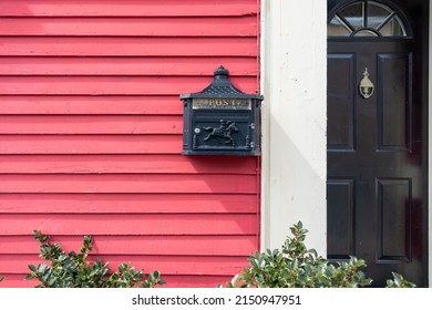 The exterior wall of a colorful red wooden clapboard siding house with a black metal mailbox, and a black glossy metal door. The postal box is affixed to the vibrant colored wall with shrubs below.