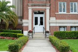 The Exterior Wall And Entrance To A Historical Brown Brick Building With Tall Columns And Concrete Steps Lead Upward To A Glass Door. There's A Palm Tree, Flowers And Shrubs On Concrete Steps.