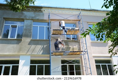 Exterior Wall Painting With A Paint Sprayer. The Building Contractors Are Renovating The Facade Of The House, Plastering And Painting On Scaffolding.