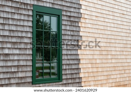 The exterior wall of a vintage building with the old section being a grey color and the new piece a light beige cedar shakes. There's a green wooden window with multiple glass panes in the old house. 