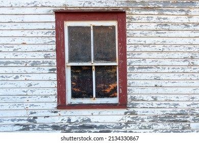 The exterior wall of a vintage white shed with a single hung closed window. The trim on the windows is a vibrant red. The white paint is peeling on the wood siding of the retro storage building. 