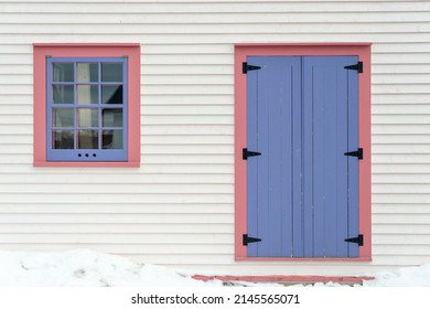 The Exterior Wall Of A White Wooden Cape Cod Clapboard Siding House With A Purple Panel Door And Vintage Glass Window, Black Metal Hinges, Pink Trim Around The Door, And White Snow On The Step. 