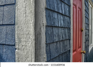 The Exterior White Wooden Corner Of An Old Textured Cedar Shake House. The Vintage Walls Are A Shiny Navy Blue Color, A White Closed Window And There's A Bright Red Door With A Brass Knob And Lock. 