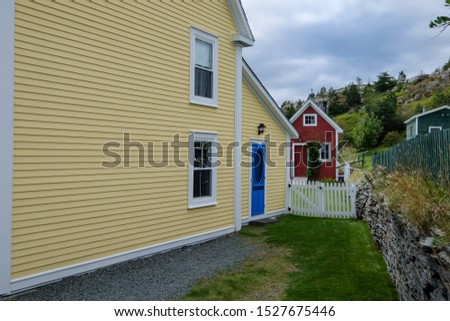 The exterior wooden wall of a historic yellow building with a blue door and white trim. There's a small red building in the rear yard of the building with a white picket fence dividing the property. 