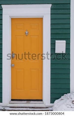 Exterior yellow metal closed door on a green building with a white metal mailbox. The ground near the door has drifts of white snow.  The building has a teal green horizontal wood clapboard wall.