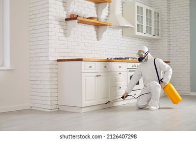 Exterminator fighting insects in house. Pest control home service guy in mask and white protective suit spraying poisonous gas or liquid from sprayer bottle on floors and cupboards in kitchen interior
