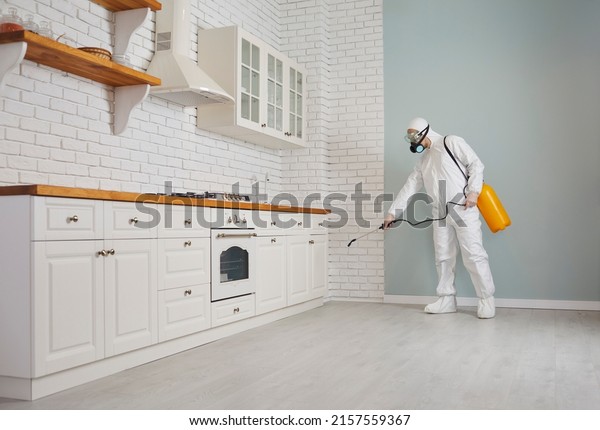 Exterminator fighting rodents, termites or\
cockroaches in the house. Pest control guy in a mask and protective\
white suit spraying poison from a sprayer bottle all over kitchen\
cupboards and\
cabinets