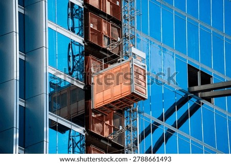 An external construction elevator is affixed to the side of a contemporary glass building, reflecting the clear sky and surrounding structures