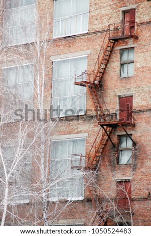 External fire escape staircase on an old brick building of factory, plant. Big windows, white birch trees grows near