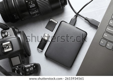 External hard disk connected to laptop, SD memory card, flash drive, camera and lens on gray textured table, flat lay