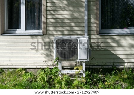 External heat pump unit positioned on ground beside wall of old wooden house, forming contrast between modern equipment and aged structure