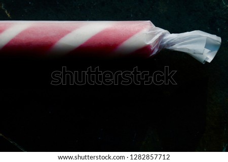 An extra large peppermint stick rolled in wax paper, lit up against a completely black background. Red and white stripes. Hard candy. Swedish candy.