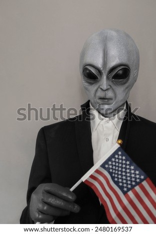 Extra Terrestrial Being, Portrait Of Alien Wearing Suit And Holding American Usa Flag on Gray Background. World UFO Day, Unidentified Flying Objects. Vertical Plane. Grey Humanoid. High quality photo