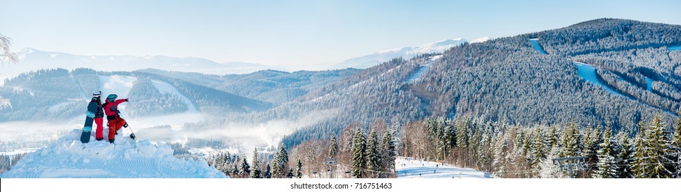 Extra wide panorama of the Carpathians mountains landscape, forests in a white haze, ski slopes, winter ski resort Bukovel. Two snowboarders enjoying, resting on top of the mountain on a sunny day