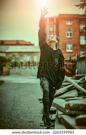 An extraordinary brutal mature man with dreadlocks and tattoos in black clothes with ethnic ornaments poses expressively on the backstreet. Rock and punk culture. 