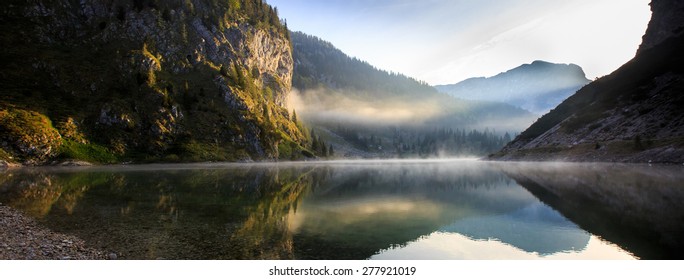 Extraordinary, fairy tale, mythological mountain lake scenery in a glacial valley, with sun shining to the rocky slope and morning mist above the lake surface with perfect reflection. 