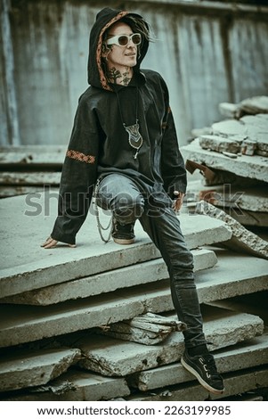 An extraordinary mature man with dreadlocks and tattoos in black clothes with ethnic ornaments poses expressively on the backstreet. Rock and punk culture. 