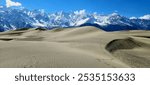 An extraordinary view of a desert landscape, where golden sands stretch out under a brilliant sun, contrasted by majestic snow-capped mountains rising in the background. This unique juxtaposition of a